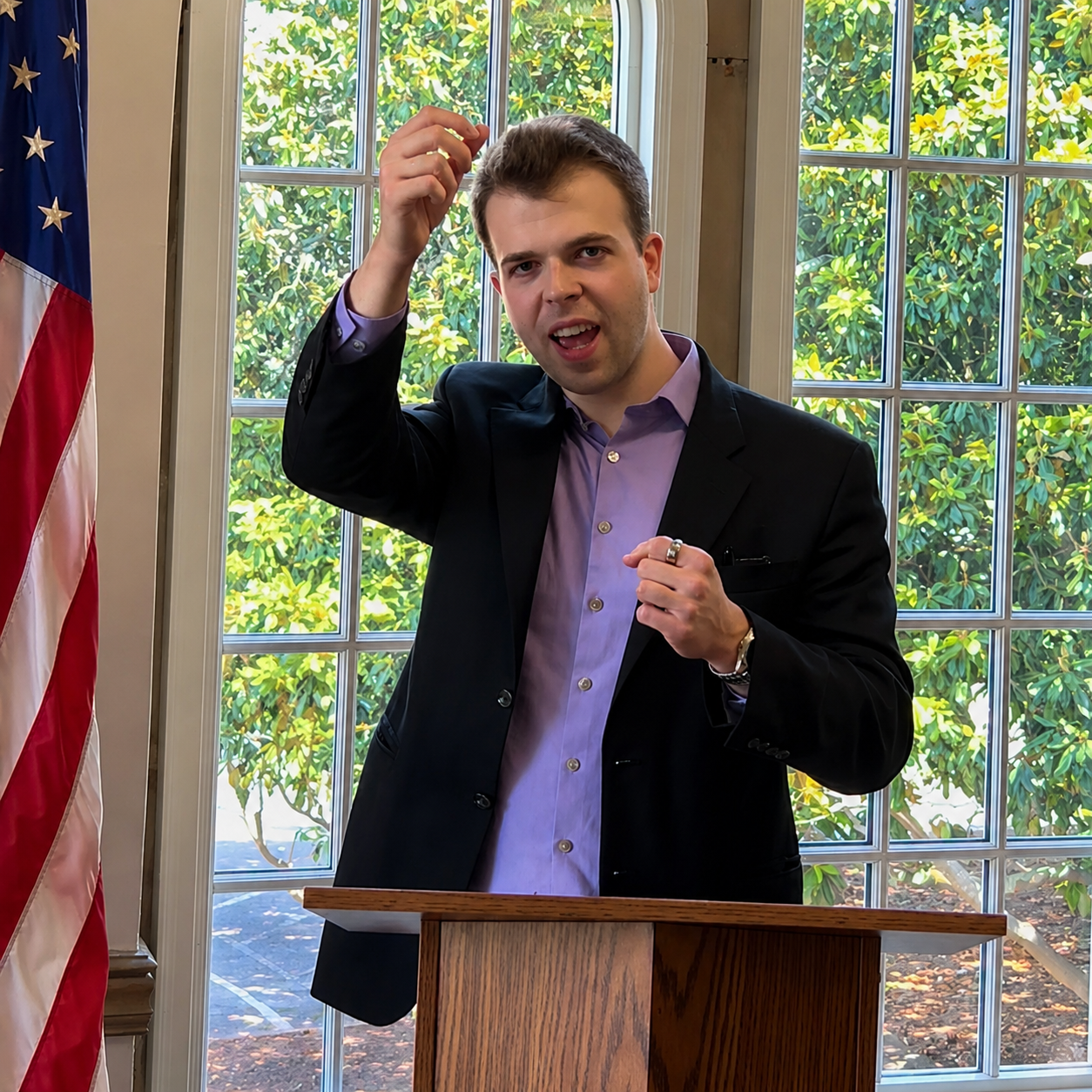 JD Hopper speaking at a podium in front of windows and an American flag